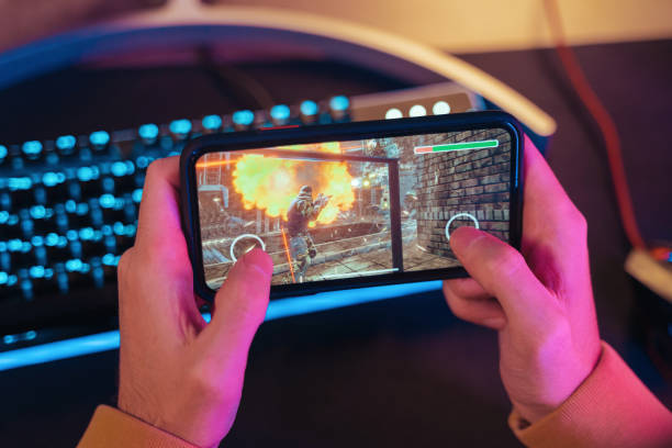 Hands of gamer playing mobile video games on his smartphone Close up of male pro gamer's hands holding smartphone with online video shooter game. Cyber sportsman playing mobile games on his cellphone. Professional keyboard on background. Top view. Neon lights gaming chair photos stock pictures, royalty-free photos & images