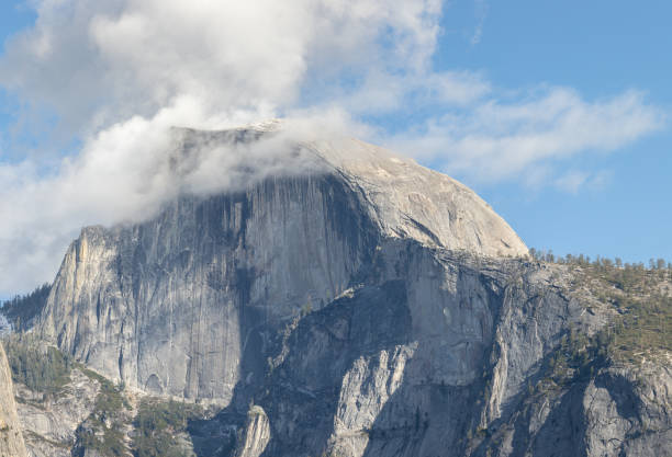 Half Dome Mountain is viewed up close. The rigid details and cracks in the rock are seen up close as fluffy white clouds pass over the mountain peak.
