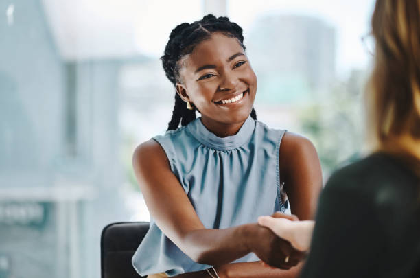 confident young african businesswoman shaking hands with a colleague in an office - gesturing interview business sitting imagens e fotografias de stock