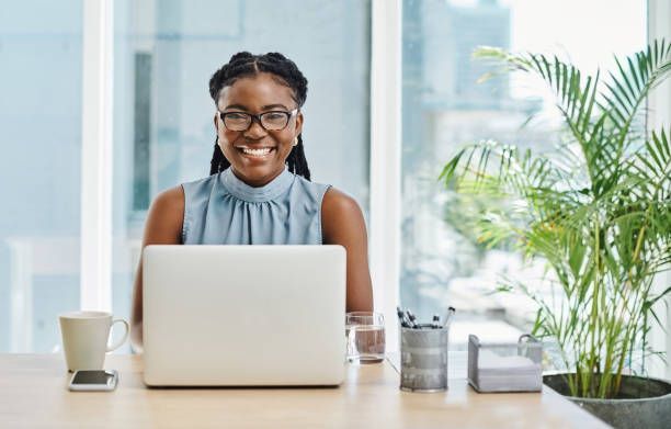 Happy african businesswoman wearing glasses and working on a laptop in an office alone stock photo