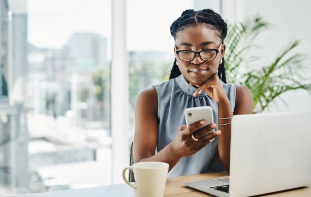 giovane donna d'affari nera che usa un cellulare mentre lavora su un laptop in un ufficio da solo - navigare in internet foto e immagini stock