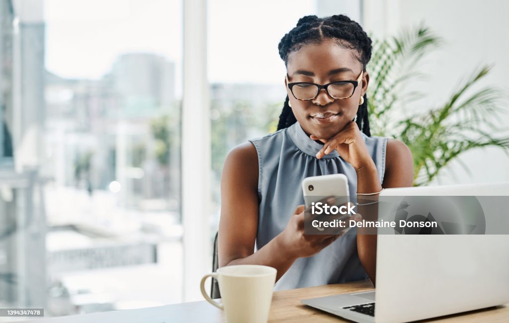 Young black businesswoman using a cellphone while working on a laptop in an office alone Women Stock Photo