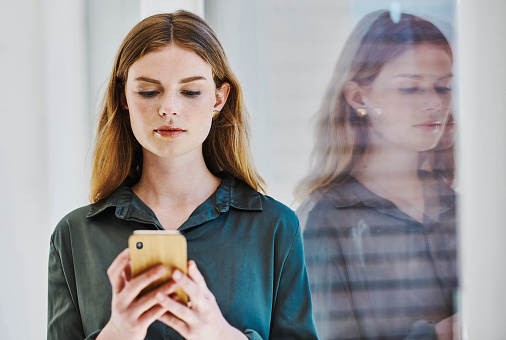 Focused young caucasian businesswoman using a cellphone while standing at a window in an office alone