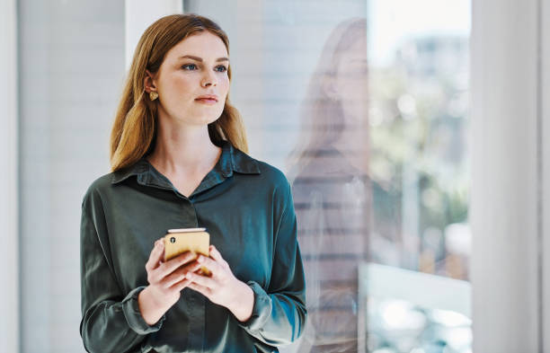 istock Young caucasian businesswoman thinking and using a cellphone while looking out the window in an office alone 1394349703