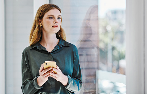 Young caucasian businesswoman thinking and using a cellphone while looking out the window in an office alone