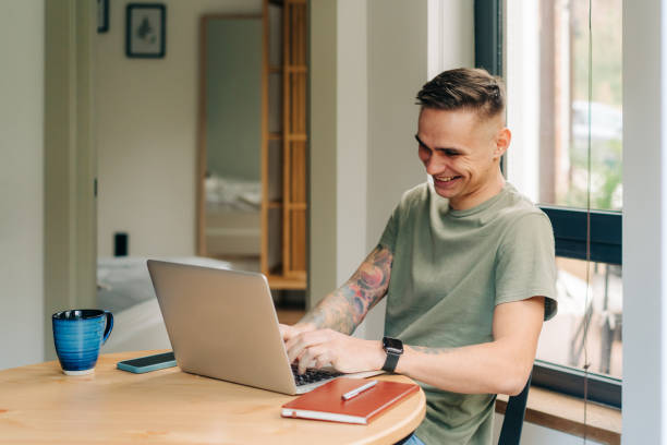 istock Cheerful laughing young man works on a laptop computer at a table at home. 1394350601