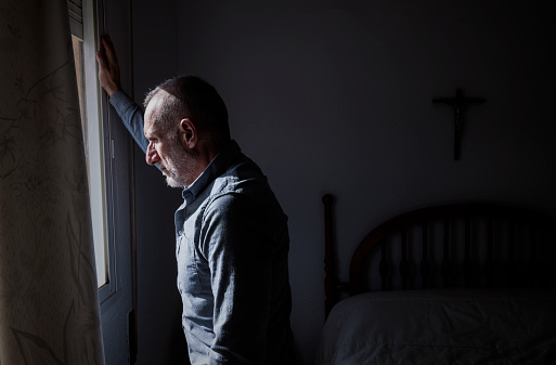 Adult man looking through window in bedroom with religious cross on wall