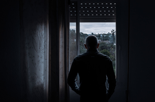 Rear view of adult man looking through window in bedroom