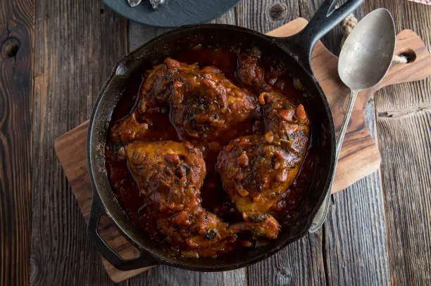 Delicious chicken dish with braised chicken legs or shanks. Cooked in a delicious and spicy tomato sauce. Served in a rustic cast iron pan on wooden table background. Overhead view