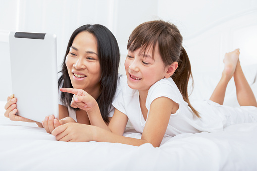 Smiling multiracial mother and daughter pointing at digital tablet laying in bedroom.