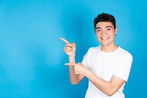 Happy, cute and smiling hispanic teenager boy pointing to copy space isolated on blue background.