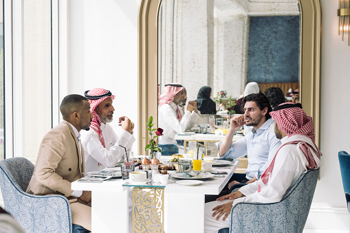 Mixed age group of Middle Eastern men in traditional and western attire exchanging ideas during a break from meetings in hotel restaurant.