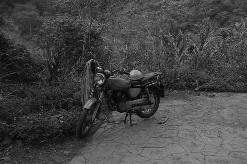 A black and white photograph of an old motorcycle found parked in the mountains with a helmet and a coat hanging  on it surrounded by tall weeds and grass.