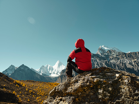 rear view of asian man sitting on top of rock with Yangmaiyong (or Jampayang in Tibetan) mountain peak in the distance in Yading, Daocheng County, Sichuan Province, China
