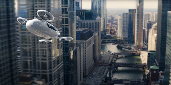 A generic electric vertical take off and landing aircraft flying high off the ground through high rise buildings through a modern urban downtown cityscape. The white eVTOL has two front and two rear rotors and is flying at speed above a river.