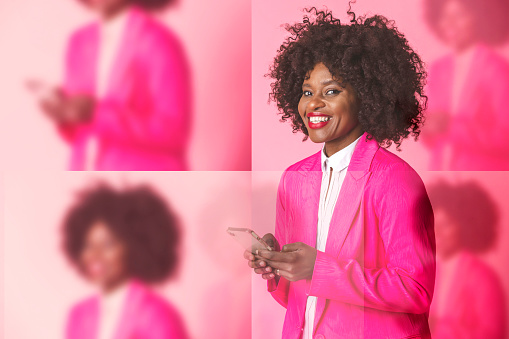 A creative portrait of a cheerful woman stood holding a mobile phone and looking into the camera. She is wearing a striking pink blazer and is standing in front of a pink background.