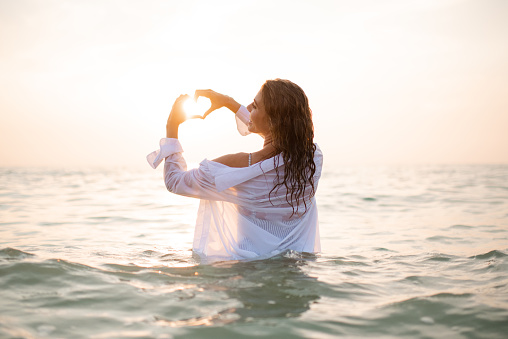 Happy beautiful woman wear wet white shirt making heart shape with hands over sunset light standing in sea. Summer season.
