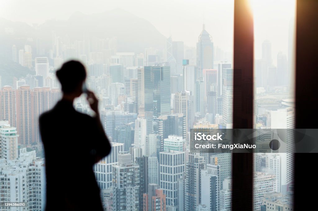 Businesswoman talking on phone from office window overlooking Hong Kong cityscape Whistleblower - Human Role Stock Photo
