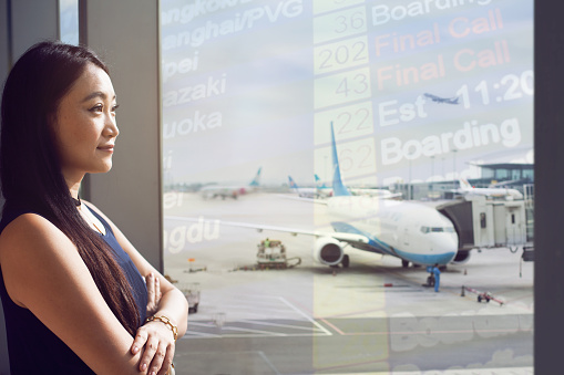 Young Chinese woman standing at airport terminal departure area looking at airplane, China