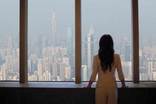 Young woman in suit looks at Shenzhen skyline from window in building