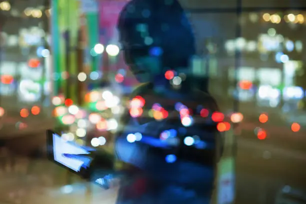 Photo of Reflection of woman in window glass at night touching a tablet computer device downtown