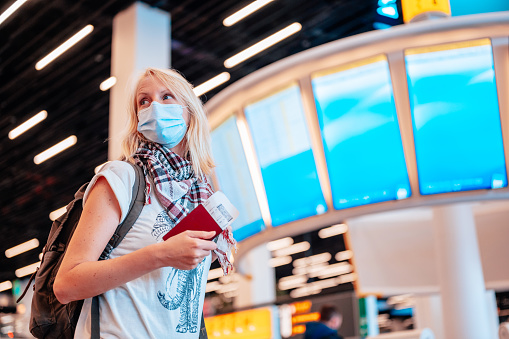 A woman at the airport holding a passport with a boarding pass in front of the flight departure board