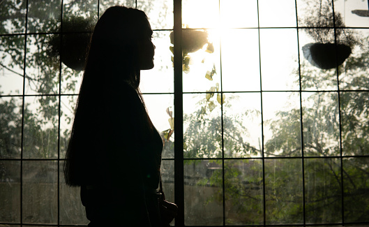 The rear view silhouette image of a happy young woman standing near the big glass window and looking at the view of the sunset through the window and thinking.