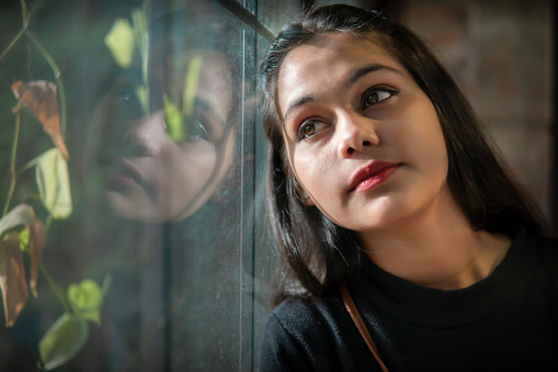 Close-up portrait of beautiful Asian, Indian pensive young woman sitting near the glass window and contemplating while looking away from the window.