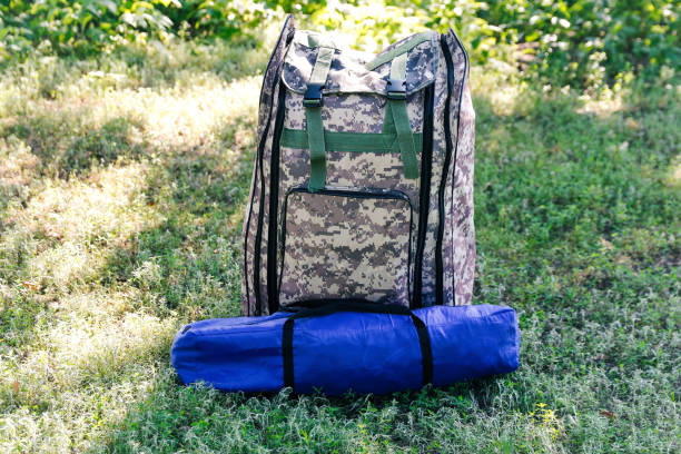 defocus military backpack and blue tent or sleeping bag. army bag on green grass background near tree. military camouflage army rucksack. vertical. tourist summer hiker. trip concept. out of focus - military canteen imagens e fotografias de stock