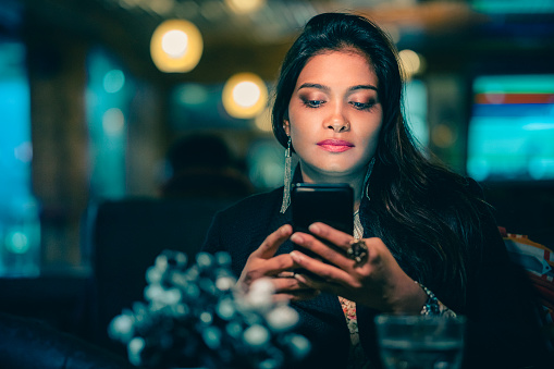 In this indoor image with copy space, an Asian/Indian serious and confident businesswoman relaxes and uses a smartphone in a restaurant after her office is closed in the evening.