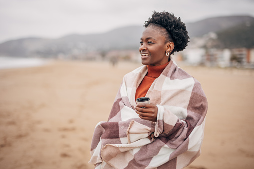 Young black women wrapped with blanket drinking coffee on the beach