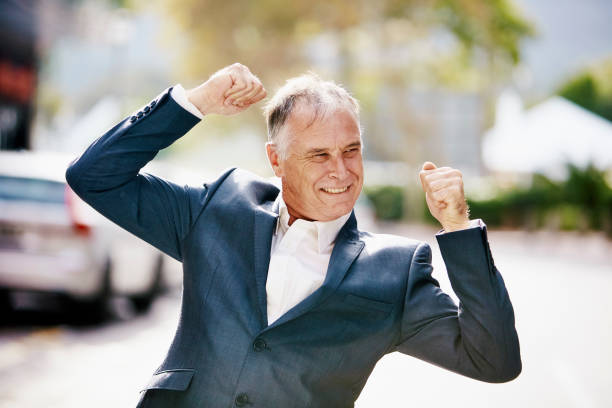 An elderly man in a suit shows a victory sign with his fingers. Gray background