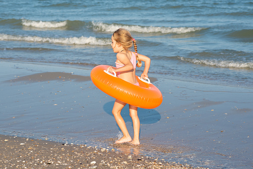 Funny little child girl running along beach with bright orange inflatable swimming ring. Vacation or holiday concept. Children girls with inflatable swimming rings having fun in blue sea water.