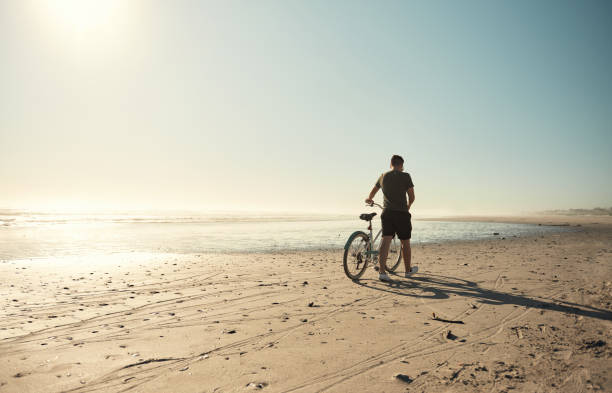 Back view of a young man walking on the beach with a bicycle
