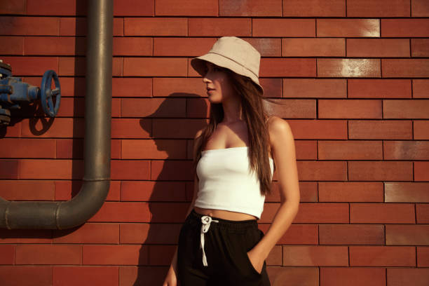 Stylish woman standing near brick wall Confident young female wearing trendy bucket hat standing with hand in pocket against wall of building in city and looking away bucket hat stock pictures, royalty-free photos & images