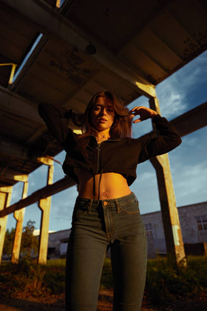 Low angle of cool young brunette in jeans and crop sweat jacket standing under grunge bridge and looking at camera in sunset light