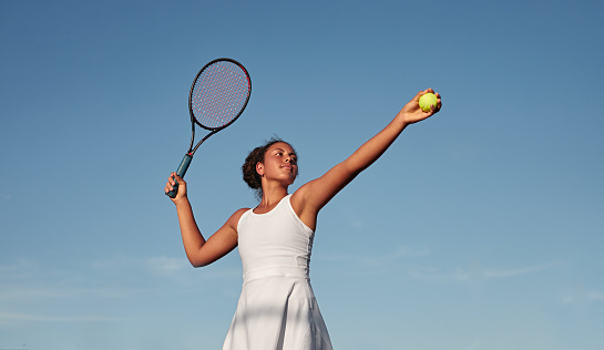 From below African American female athlete in white dress preparing to hit ball with racket against blue sky while playing tennis in daytime