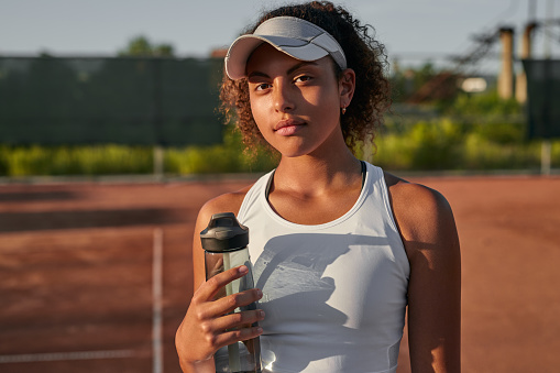 Young African American female athlete in sportswear and with bottle of water standing on sports ground in sunset in summer and looking at camera