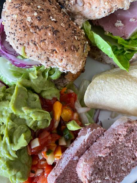 Stock photo showing close-up, elevated view of blue rimmed plate containing ham and salad granary roll sandwiches. The salad includes plate, lettuce, ham, grated carrot, chopped cherry tomatoes and red onion rings. imagen de fotograma completo del plato que contiene sándwiches de rollo de granero de ensalada de jamón, lechuga, jamón y rollos de aros de cebolla roja servidos con guacamole, salsa de tomate, rodajas de carne en conserva y patatas fritas, vista elevad