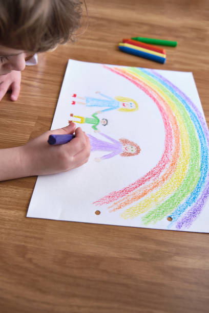 From above crop kid creating picture of LGBT family under rainbow with crayons while lying on floor on weekend day at home