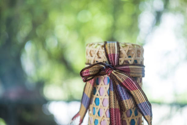 A sticky rice container, made of bamboo strips woven into a pattern and shaped of a cylinder. This basketry has two parts, the container and the cover tying together by the red rope.