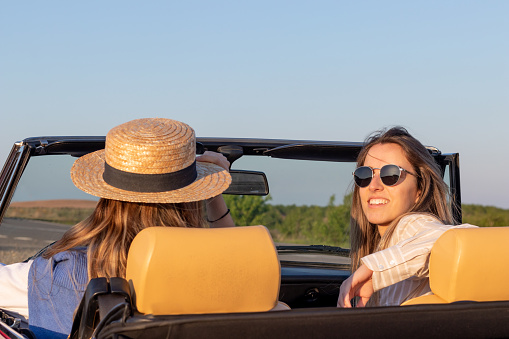 Close up of two young women driving a convertible car while looking at camera on a sunny and blue summer day. Wearing straw hat and sunglasses