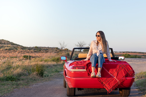 Young beautiful girl sitting on a red classic convertible car enjoying the sunset on a remote road in the countryside in summer