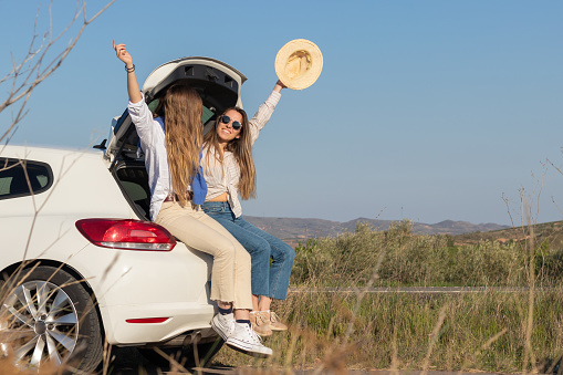 Two friends having a good time during spring accations sitting in the back of the car while raising their arms and hat. with a landscape in the background