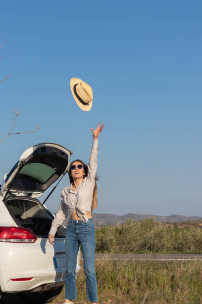 vertical picture of teenager on sunglasses having fun throwing her straw summer hat in the air while in a road trip - grass area field air sky imagens e fotografias de stock