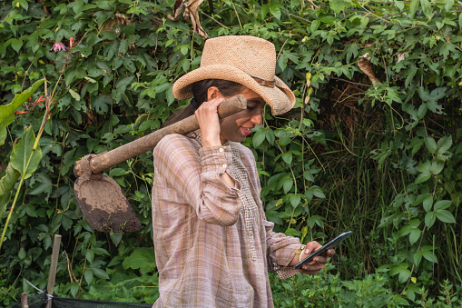 Peasant woman holding a hoe over her shoulder looking her smartphone in a orchard
