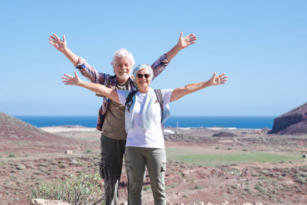 feliz pareja de ancianos activos en excursión de montaña con los brazos abiertos disfrutando de un estilo de vida saludable y un día soleado. dos ancianos sonrientes con mochila durante las vacaciones, horizonte sobre el agua. - horizon over water environment vacations nature fotografías e imágenes de stock