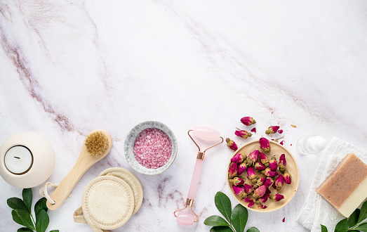 flat lay with organic sea salt with dried rose flowers, soap, candle, brush and gua sha on a pink marble background. The concept of a natural spa product. Top view and copy space.