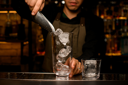 front view of the transparent mixing cup into which the hand of male bartender pours ice cubes using scoop. Glass with ice nearby on the bar counter