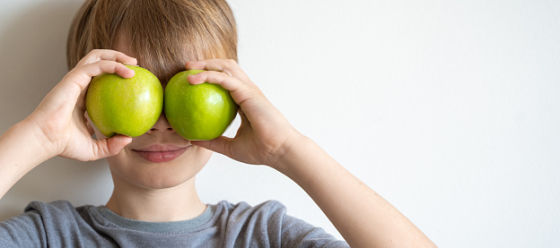 Cute child in grey t-shirt holding green apples in front of his eyes over white background. Concept of healthy nutrition.